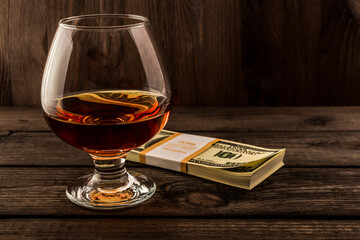 Glass of brandy with pack of dollars on an old wooden table. Focus on the glass of brandy