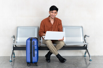 Fototapeta na wymiar Latin man sitting at station with suitcase and working with laptop