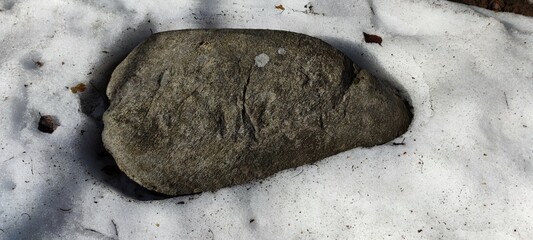 Large oval granite stone. In the midst of melting white snow under the sun lies a large granite...