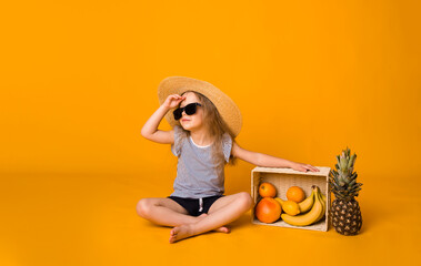 a beautiful little girl in a straw hat and sunglasses sits with a basket of fruit on a yellow background and looks away.