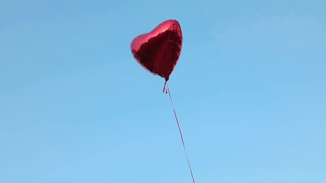 A red foil balloon in the shape of a heart filled with helium flies against the backdrop of a cloudless sky. Concept for love, Valentine's day, relationship. Full HD video footage