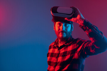 bearded man with tattoos raising his virtual reality glasses above his eyes. Studio portrait with colored lights of person using modern technology.