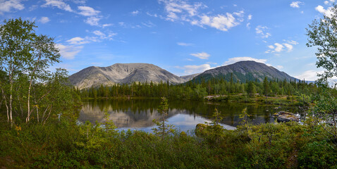 Panorama of a mountain lake surrounded by a spruce forest against the background of a mountain range and a blue sky with white clouds