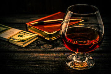 Glass of brandy and leather notebook with golden pen and pack of dollars on an old wooden table. Angle view, focus on the glass of brandy