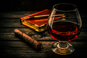 Glass of brandy and cuban cigar with leather notebook and golden pen on an old wooden table. Angle...