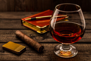Glass of brandy and cuban cigar with leather notebook and golden pen on an old wooden table. Angle view