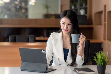 Beautiful Asian businesswoman holding a coffee cup and working on the tablet at office.