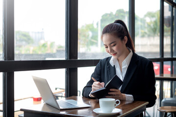 Charming Asian business woman taking notes by the window with a laptop placed at the table. Working concept.