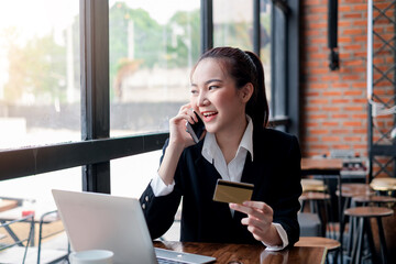 Asian businesswoman sitting by the window talking on the phone looking outside holding a credit card laptop empty at the office desk.