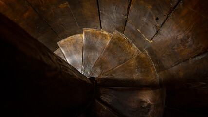An old wooden spiral staircase leads down to a dark and narrow room.