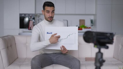 Portrait of confident young Middle Eastern financial advisor pointing at graph talking at video camera in home office. Handsome bearded man explaining business for video blog indoors