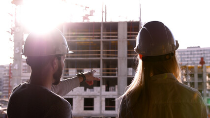 Young civil engineer with a beard in a white helmet and a beautiful female builder in a white construction helmet stand at a construction site and talk against the background of a house under construc