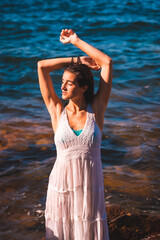 Fototapeta na wymiar A young Caucasian woman enjoying with her eyes closed in a white dress and wet hair sitting on a rock in the sea, lifestyle