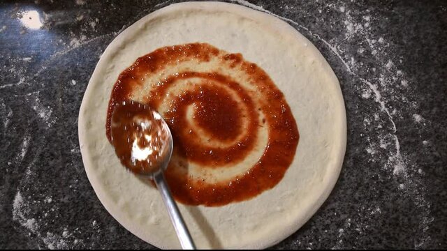 Pizza cooking process. A chief applying tomato ketchup with metal spoon on pizza base. Top view of the base for pizza is added natural tomato sauce.
