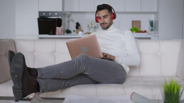 Wide shot of wealthy rich confident young handsome man in headphones smiling typing on laptop keyboard. Portrait of successful Middle Eastern manager messaging online in home office