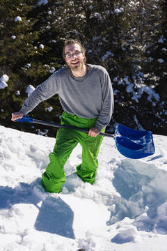 Full Figure Portrait From Above Of A Young Man Knee Deep In The Fresh Snow On A Roof, Posing For Camera With A Big Smile An A Blue Shovel In His Hands