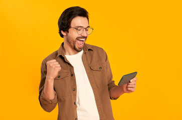 Young bearded man with glasses looks at the smartphone screen and shows the gesture yes, winner,...