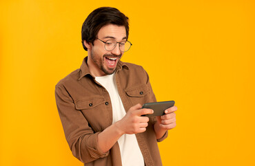 Young bearded man in glasses plays games enthusiastically on the smartphone isolated on orange...