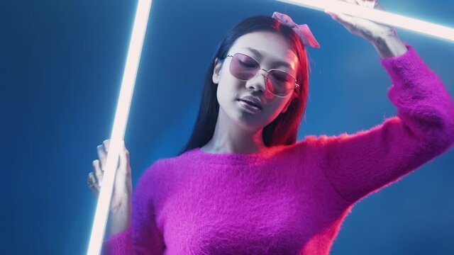 Neon light people. Cyberpunk fashion. Futuristic beauty. Glamour Asian model girl in glasses pink sweater in red color glow posing with white LED lamp isolated on blue.