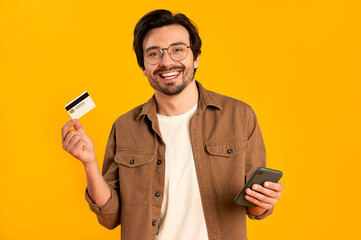 Young bearded man in glasses holds credit card and smartphone isolated on orange background. Online...