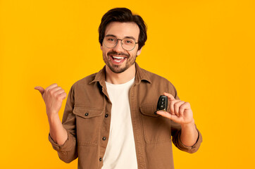 Young bearded man in glasses holds keys remote control from a new car isolated on orange...