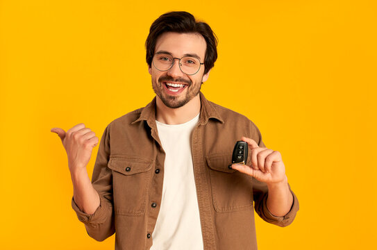 Young bearded man in glasses holds keys remote control from a new car isolated on orange background. Purchase, car rental.