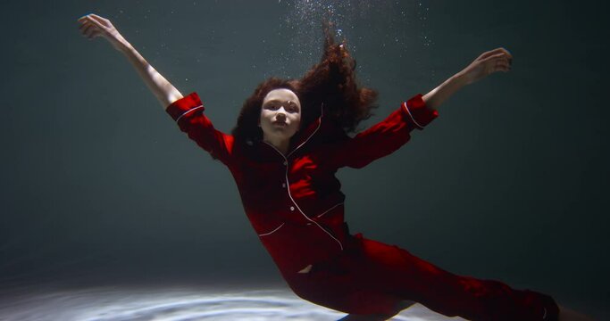 Ethereal cinematic underwater portrait of young beautiful woman in red pyjamas looking at camera, arms open slow motion.