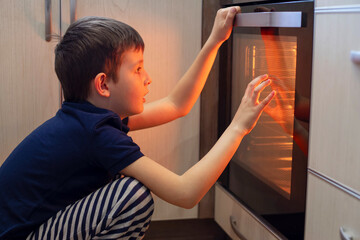 Fototapeta na wymiar A child is sitting near the oven in the kitchen and waiting. Curious boy is watching through the glass of kitchen oven. Baking pizza, muffins , cupcakes or cookies