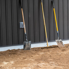 Dirty, used shovels and a rake stand upright on the dirt and lean against the board and batton...