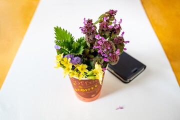 Bouquet of spring flowers in coffee crucible and smartphone on empty table.