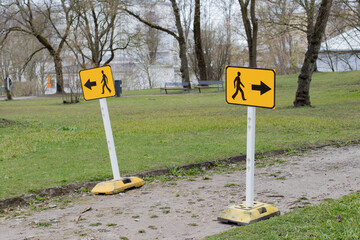 Two detour traffic signs with arrows to opposite directions for pedestrians on a muddy sidewalk for construction work