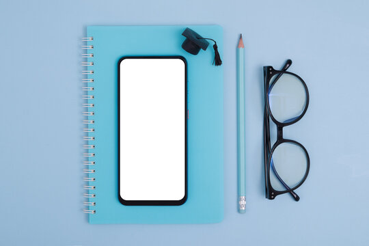 Top View Of Stationery, Glasses With Phone Mockup And Graduate Cap On Blue Background. Monochrome Online Education Concept, Flat Lay Knolling