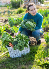 Portrait of young female gardener sitting with basket full of freshly harvested greens in vegetables garden