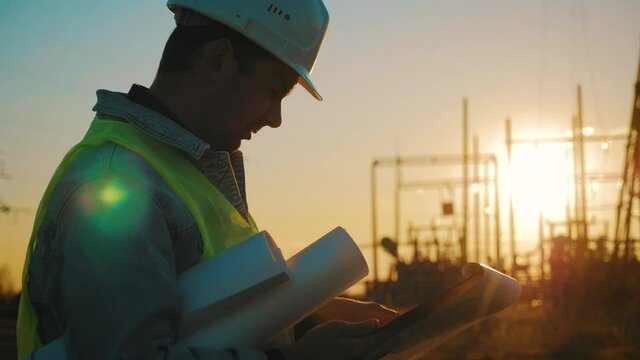 Electricity Work Concept. Silhouette Of Engineer In Helmet Standing On Field With Electricity Towers. Electrical Engineer With High Voltage Electricity Pylon At Sunset. Voltage Tower Support.