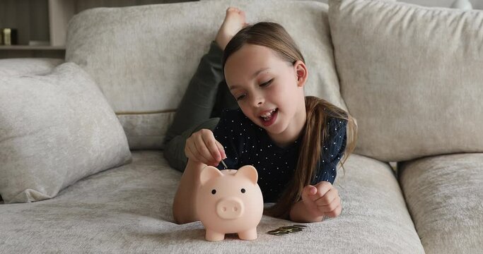 Beautiful 7s girl lying on sofa counts and dropping coins into piggy bank. Save money, pocket cash for future needs, education, purchases, learn kid be thrifty, economy skills from childhood concept