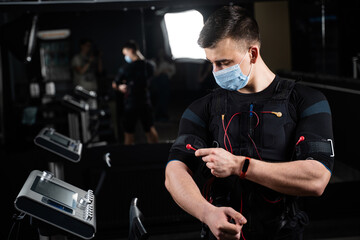 Man in EMS suit and medical mask in gym. Protection from coronavirus covid-19. Sport training in...