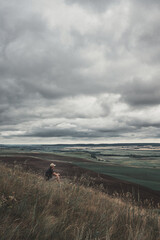 Obraz premium Back view of an unrecognizable man sitting on a hill against a gloomy cloudy sky. A lonely man on the side of the mountain. Rainy gray clouds over the valley