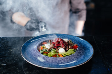 A professional chef serves a freshly prepared salad of tomato and veal greens under a glass hood with thick smoke. Beautiful smoky serving in the restaurant.