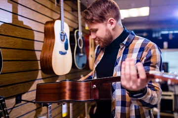 Wall murals Music store redhaired bearded handsome man in brown plaid casual shirt choosing a guitar in a music store