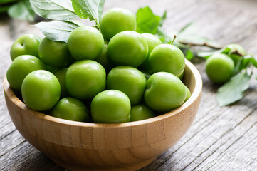Close up fresh green plums or greengage in bowl on wooden background, heap of summer fruits concept