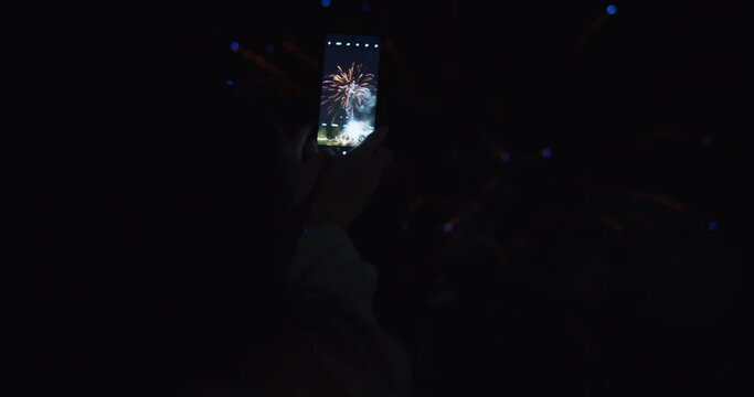 A woman taking a picture of colorful fireworks in the night sky with her smartphone. 