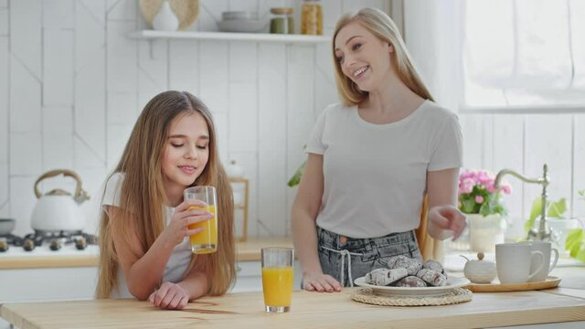 Adult beautiful blonde mother 35s woman puts on table plate with homemade chocolate cookies looks with tenderness at beloved daughter child girl drinking healthy orange juice hug baby kiss on forehead