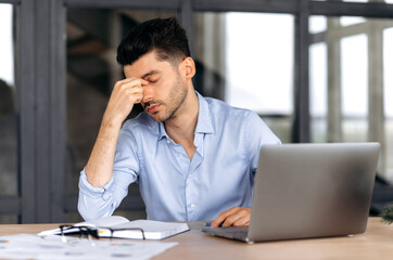 Obraz premium Headache and migraine. Exhausted caucasian young adult man, manager, freelancer or office employee, tired of works, in stress, took a break from work, need a rest, massages eyes, have a headache