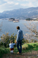 Dad holds the hand of a little girl with a soft toy, standing on the coast against the background of the sea, mountains and Sveti Stefan island. Back view.