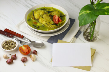 Pindang Ikan Patin or Catfish vegetables Soup , Indonesian food popular from Palembang. Served on ceramic bowl garnished with tomato slices. Blank paper and envelope for text. White marble background.