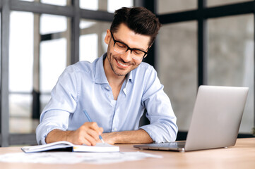Fototapeta na wymiar Smart successful caucasian man with glasses, sitting at the workplace, using a laptop, taking notes, studying or working online, listening to a webinar, gaining knowledge, smiling