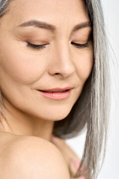 Mature older mid aged Asian woman of 50s with grey hair looking at her perfect skin. Advertising of spa procedures for women in menopause. Skincare, cosmetology, dermatology ads. Close up portrait.