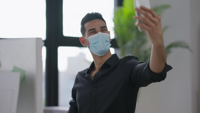 Cheerful young man in Covid-19 face mask taking selfie on smartphone in home office on sunny day. Portrait of smiling joyful positive Middle Eastern manager enjoying remote working. Lifestyle