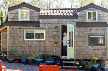 Friendly mature woman standing at front door of her charming little wooden tiny home or house - obrazy, fototapety, plakaty