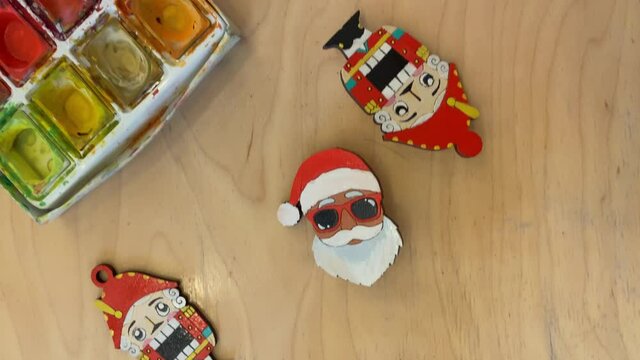 Plywood pins in form of Nutcracker and Santa Claus in hat and glasses. Painted figurines and aquarelle watercolors on wooden table. Close-up of artist's workspace.